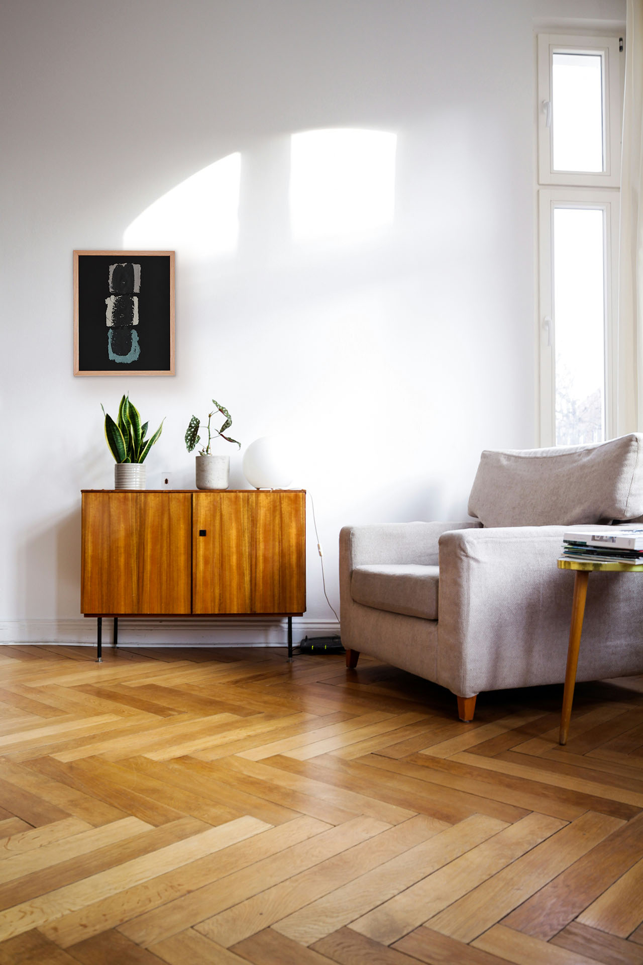 Salón de estilo mid-century con suelos de madera en espiga y la obra "Deconstruyendo III" expuesta sobre un aparador de madera vintage. Cuadro abstracto con tres formas orgánicas superpuestas en tonos crema, gris y azul suave sobre fondo negro.