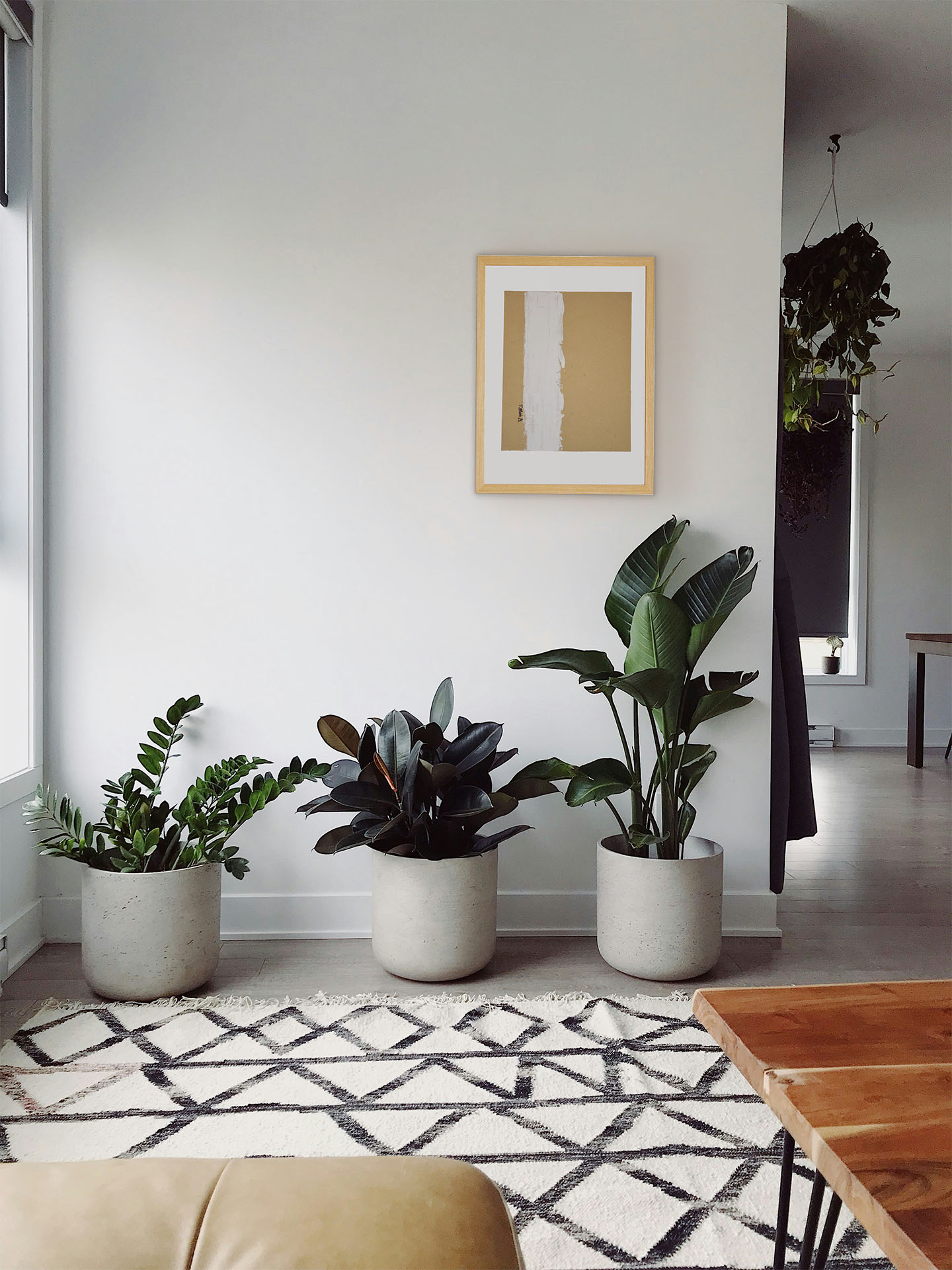 Vista de salón minimalista con tres plantas grandes en macetas de cemento. En la pared, la obra "Mi realidad I" de La Oculta Gallery, enmarcada en madera clara, muestra una composición abstracta ocre y blanca. El espacio cuenta con una alfombra geométrica y luz natural.